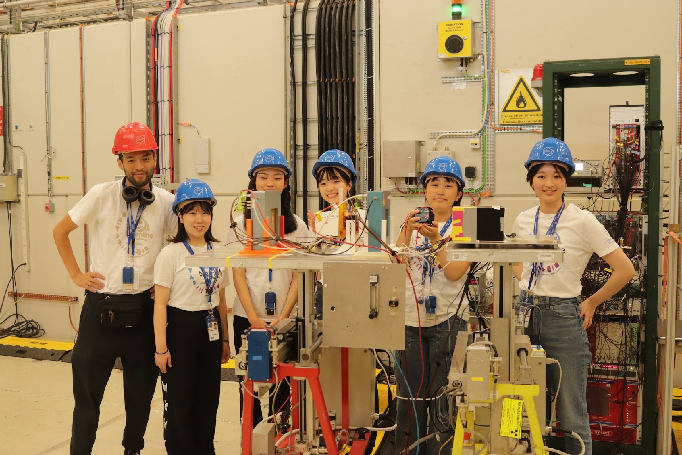 CERNのビームラインで実験装置の前に並ぶヘルメット姿の学生チームと指導者